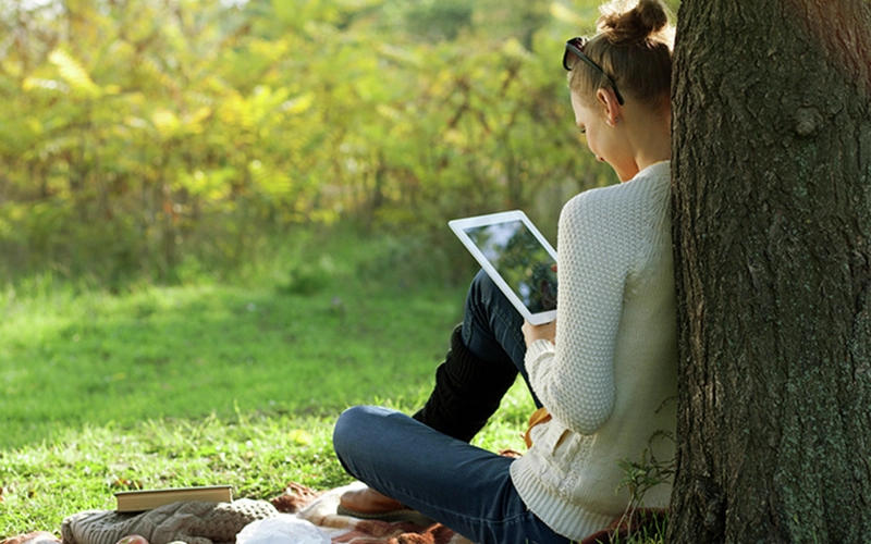 Žena sedí venku u stromu a čte na tabletu