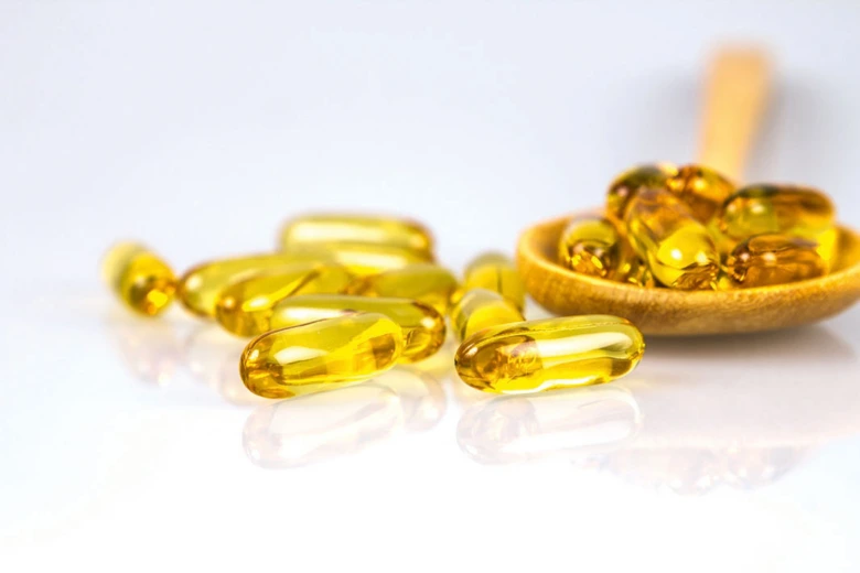 Žluté kapsle s vitamínem D na bílém pozadí vedle dřevěné lžičky.