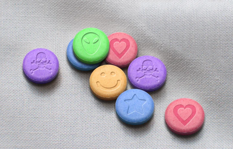 Kolorované tablety MDMA s různými symboly rozestavené na bílém povrchu.