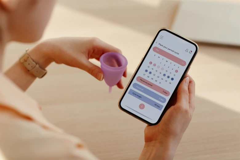 Žena drží menstruační šálíček a aplikaci pro sledování cyklu.