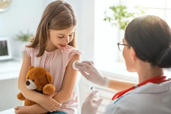 Dívka s medvídkem dostává očkování od zdravotní sestry.