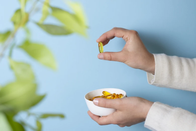 Ruce drží bílou misku s kapslemi vitamínů na modrém pozadí.