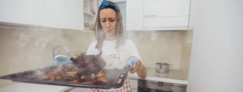 Žena drží kouřící plech s popáleným jídlem z trouby.