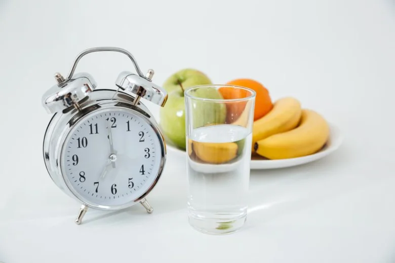 Stříbrní budík vedle sklenice vody a talíře s ovocem.