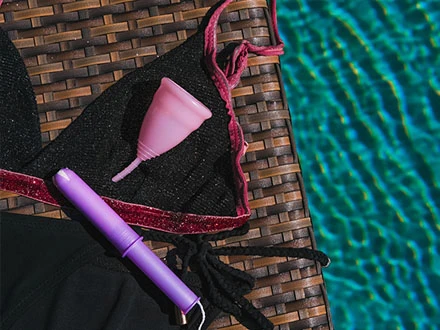 Růžová menstruační šálka a fialový tampón vedle plavek.