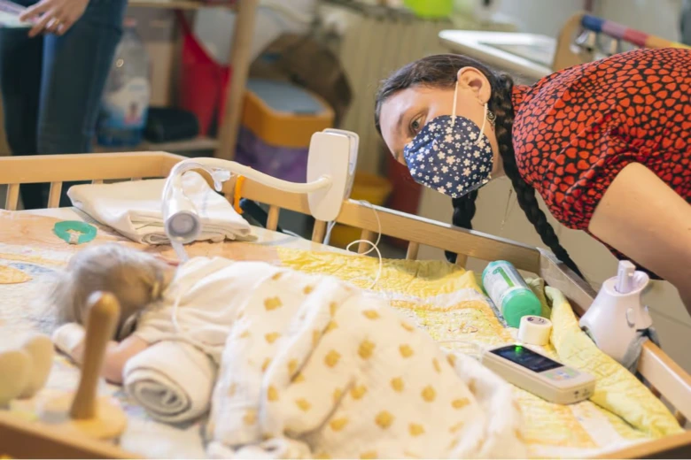 Žena v roušce se dívá na dítě ležící na lůžku s lékařským vybavením.